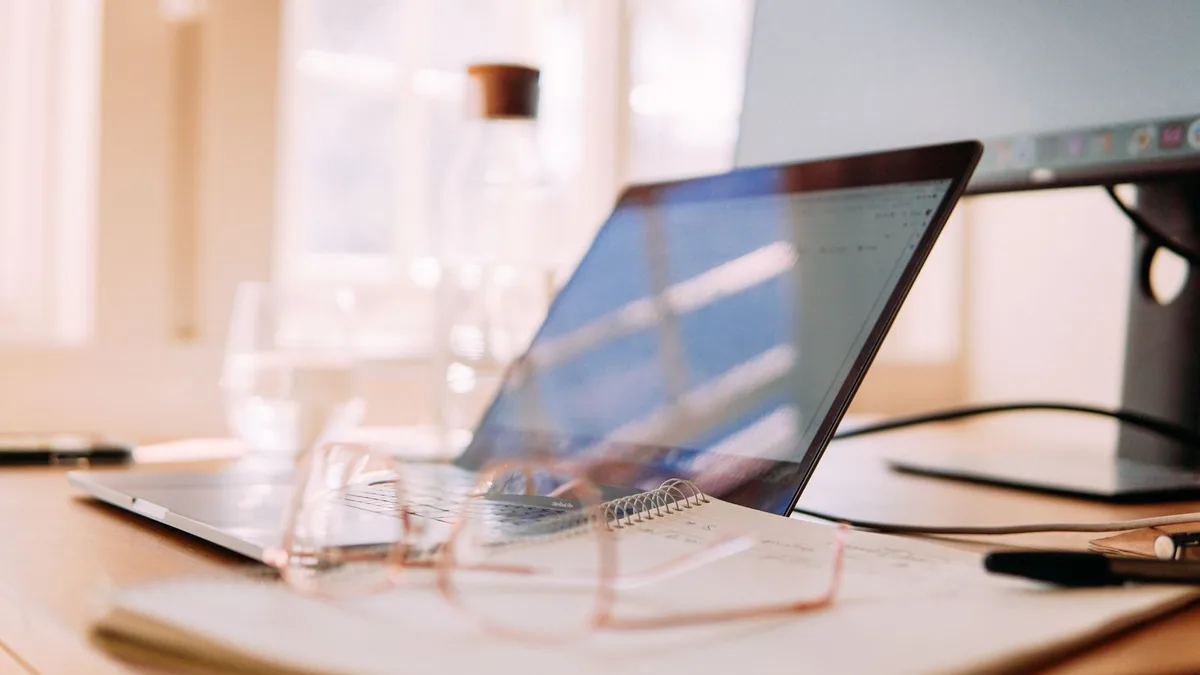 Open laptop on a light wooden desk by a window, with a spiral notebook, a pair of glasses and a pen in front of it