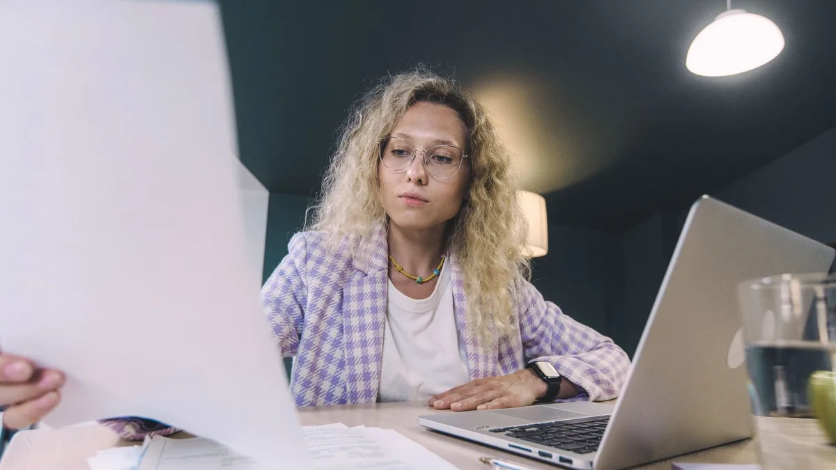 Vrouw met krullend haar en een bril leest aan haar bureau een vel papier dat ze omhoog houdt, naast een opengeklapte laptop en losse papieren