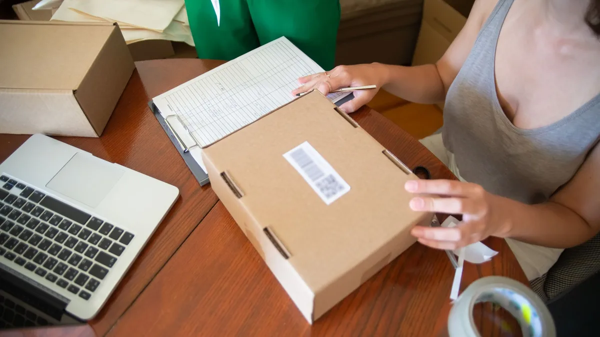 Twee handen die een kartonnen verzenddoos met verzendlabel dichtplakken op een houten tafel, met een opengeklapte laptop en een klembord ernaast